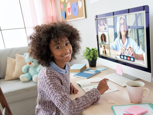 Cute small african kid girl looking at camera during social distance online class on video conference call with teacher, tutor and school children group learning at home. Elearning class on computer.